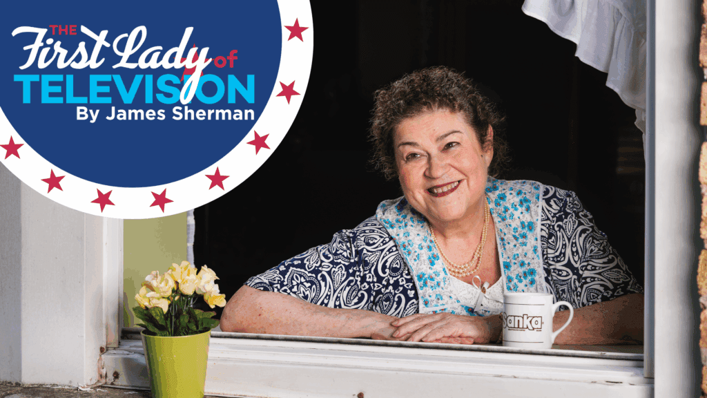 Actress Cindy Gold as Gertrude Berg posed in front of a window dressed with white curtains. a potted plant rests on the white windowsill. She holds a white coffee cup with the Sanka logo on it.