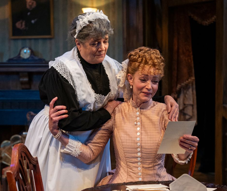 Bella is seated, reading a letter. Elizabeth stands beside her, hands comfortingly on Bella's shoulders as she reads.