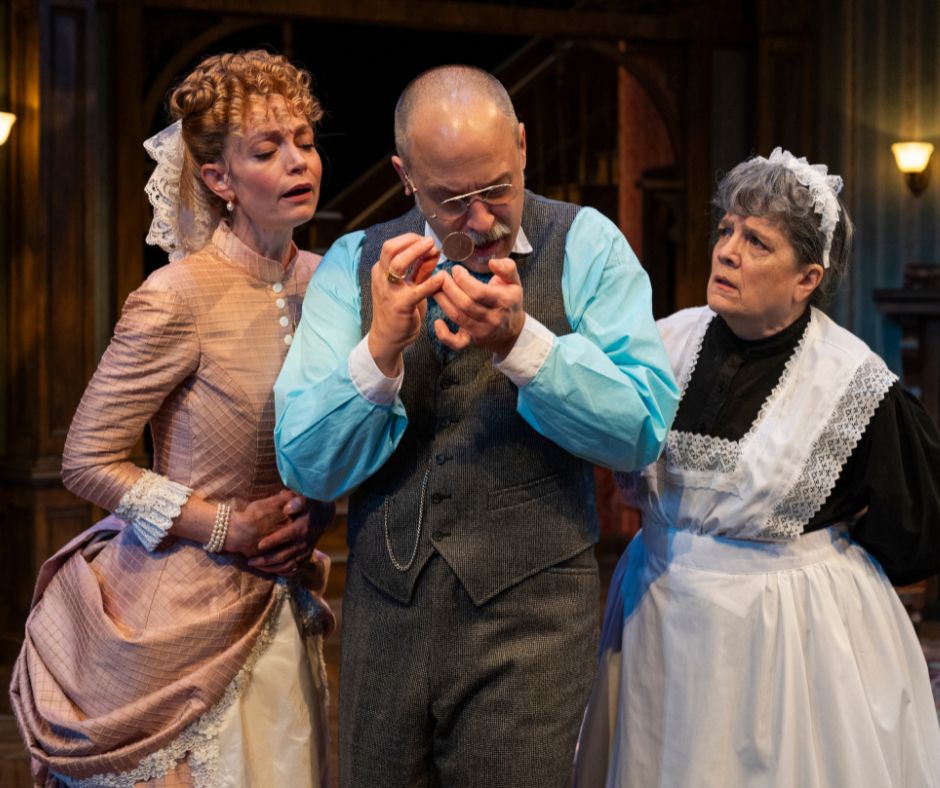 Sergeant Rough examines a small broach with a magnifying glass as Bella and Elizabeth stand on either side, looking over his shoulder.