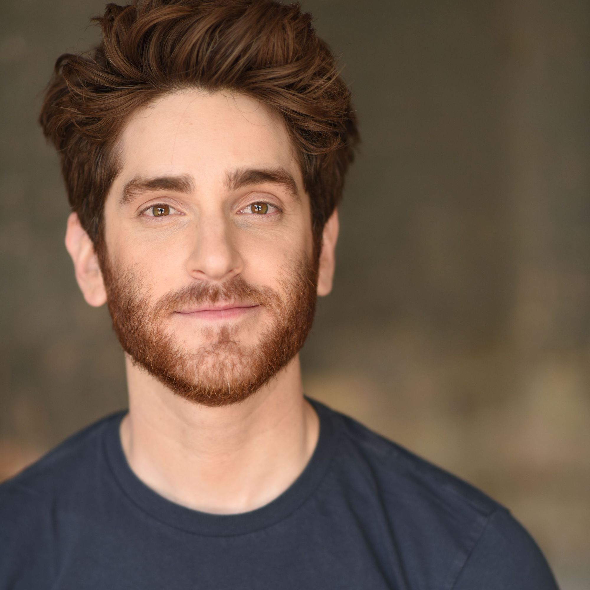actor garrett Lutz, a man with red hair, mustache, and a beard in a gray shirt pictured from the shoulders up in front of a brown backdrop