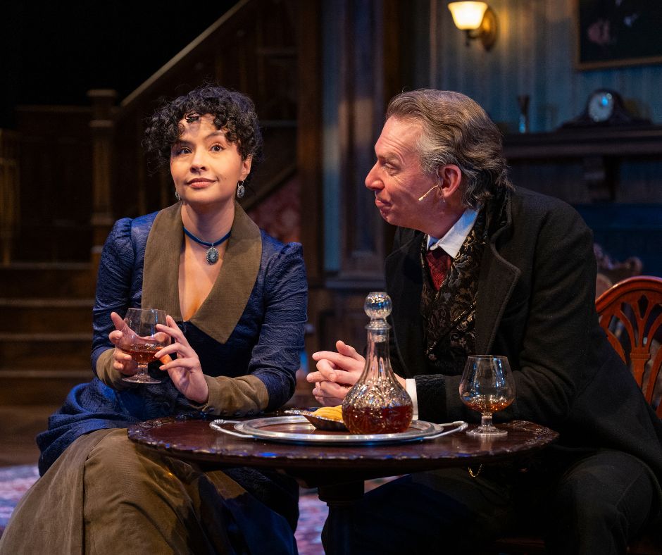 Nancy and Jack are seated side by side at a table with glasses of brandy in hand. Nancy looks outward, a small smile on her face and Jack looks at her intently.