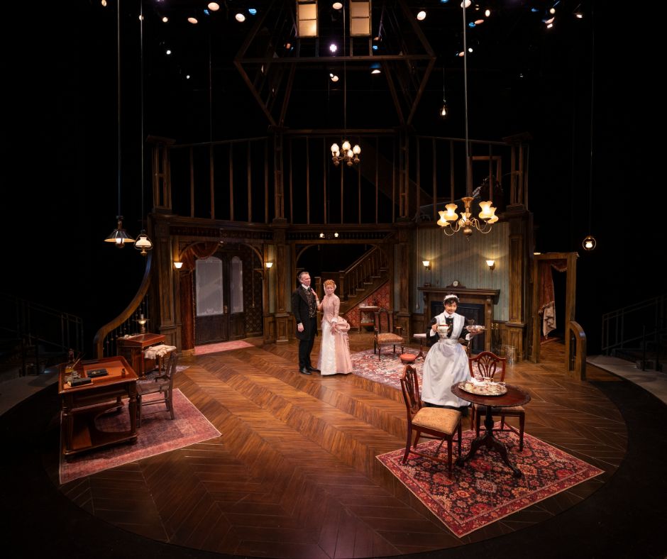 A wide shot of the set, a three story victorian house with a warm wood floor decorated with lush-colored rugs and lit by various gas lamps. Bella and Jack stand center stage, and Nancy is slightly downstage setting a tea set down on a table.