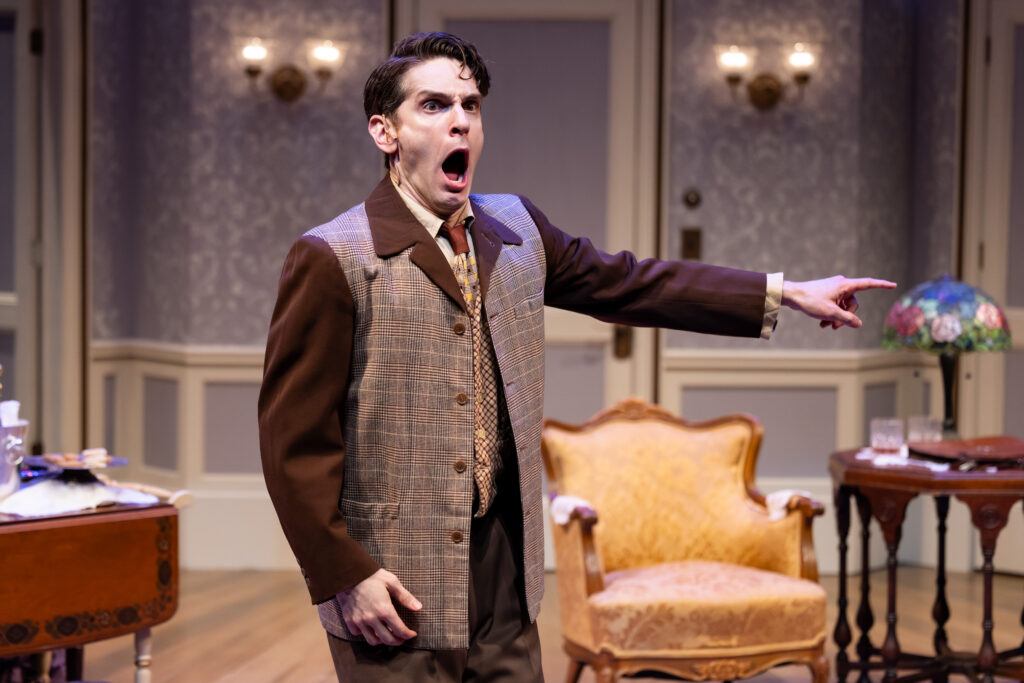 Actor Actors Garrett Lutz in a brown suit, standing with his mouth open, pointing with his arm fully extended.