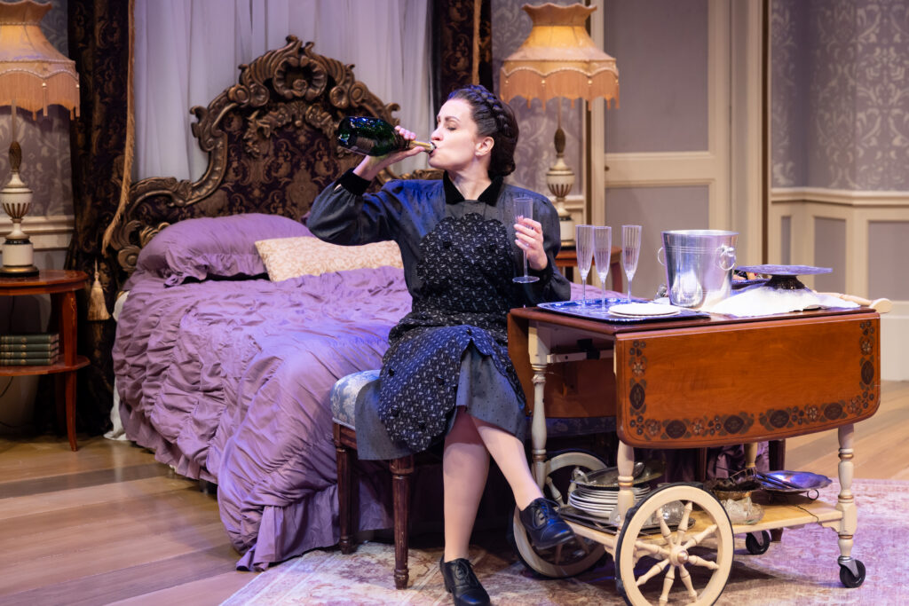 Actress Erin Noel Grennan seated on the edge of a lush purple bed, drinking champagne from a bottle.