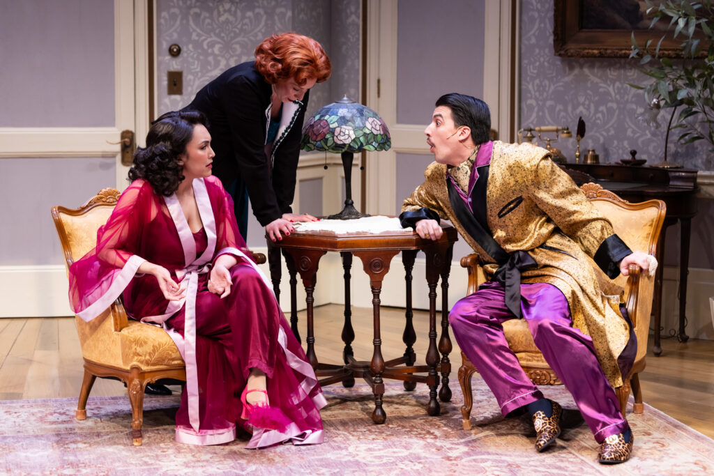 Actress Katy Sullivan standing upstage from Actress Aja Alcazar and actor Andrés Enriquez, both seated in arm chairs