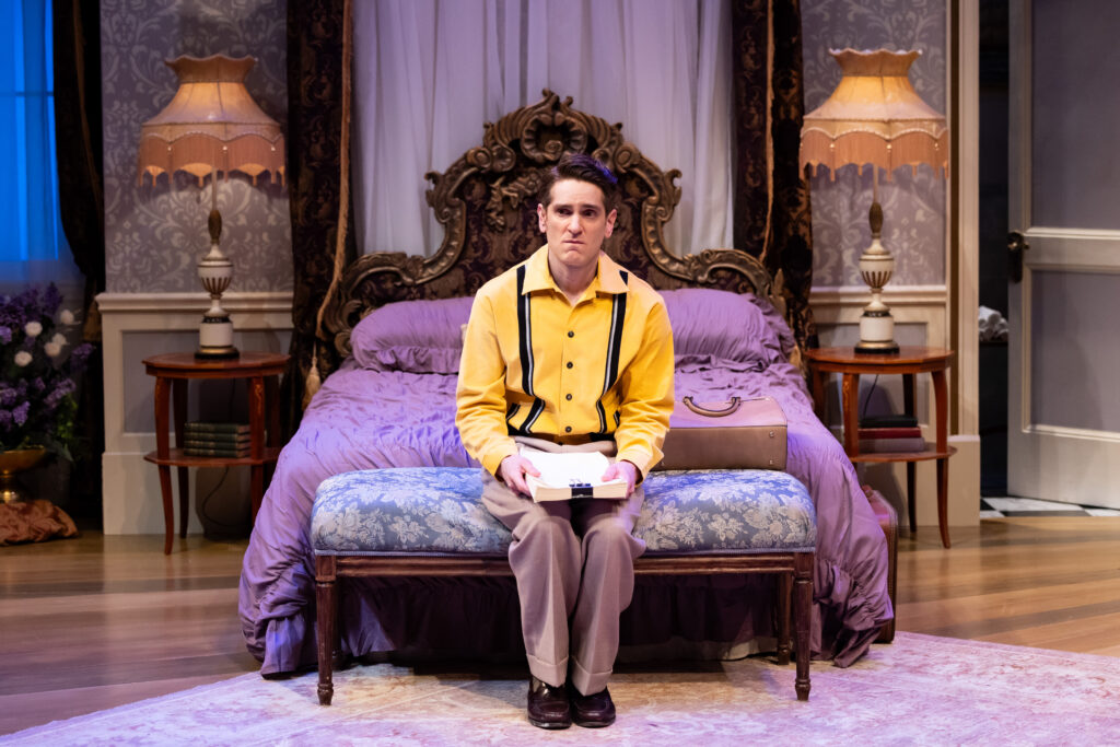 Actor Garrett Lutz at Oliver Adams, in a yellow shirt and brown pants seated on a lush purple bed