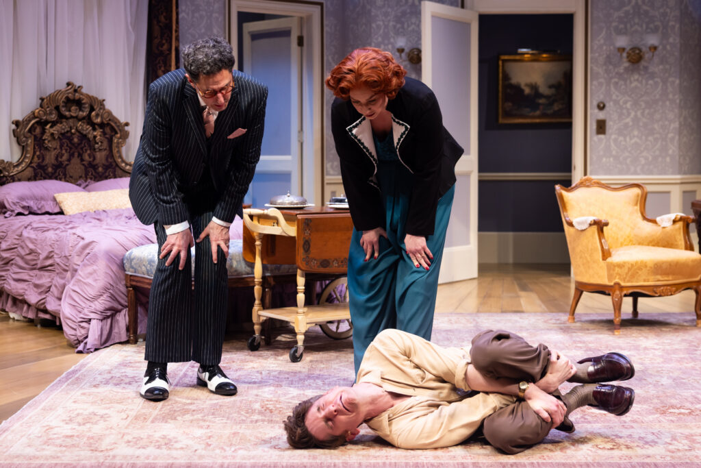 Actor Garrett Lutz in the fetal position on the floor as actors Sean Fortunato and Katy Sullivan stand over him, bent over with their hands on their knees.