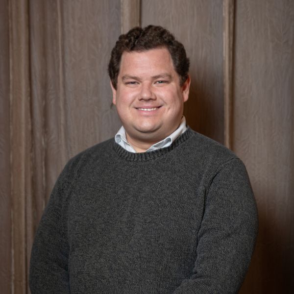 Brian Scully, a man with short brown hair in a grey sweater with a white collared shirt beneath, smiling in front of a wood panel background.