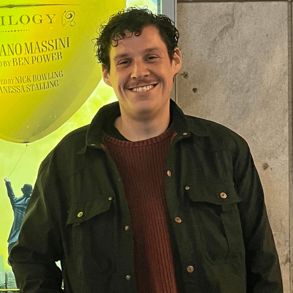 Devan Castle in a black jacket over a maroon shirt, posed and smiling in front of a musical theatre poster