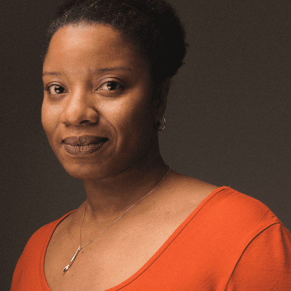 Actress Elana Elyce in an orange shirt, facing the camera as she stands in front of a dark gray backdrop