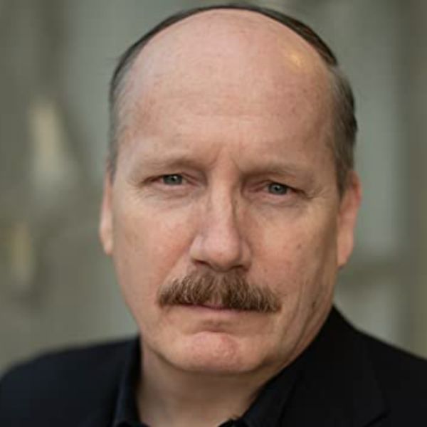 Actor Guy Van Swearingen in a dark jacket posed in front of a light gray backdrop