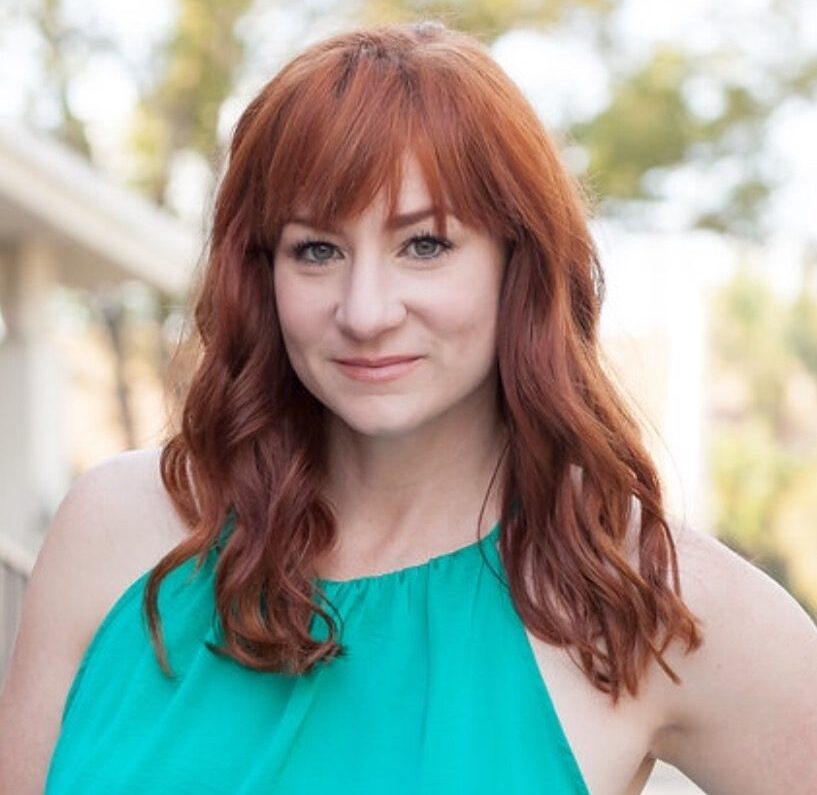 Actress Katy Sullivan, a woman with long red hair in a turquoise shirt, posed in front of an out of focus backdrop dressed with greenery. She is pictured from the sternum up.
