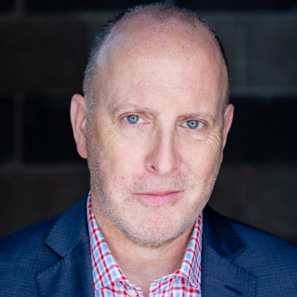 Actor Joe Dempsy in a checkered shirt and a blue suit coat standing in front of a grey backdrop
