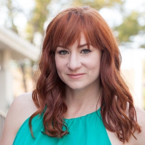 Actress Katy Sullivan, a woman with long red hair in a turquoise shirt, posed in front of an out of focus backdrop dressed with greenery. She is pictured from the sternum up.