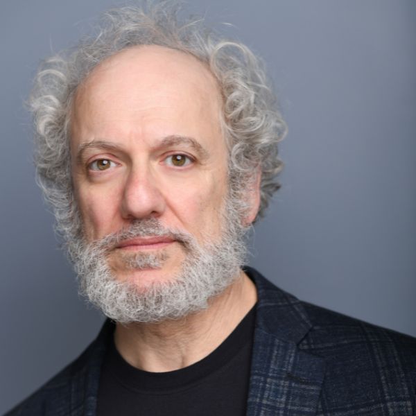 Actor Mark David Kaplan in a dark blue jacket in front of a gray backdrop