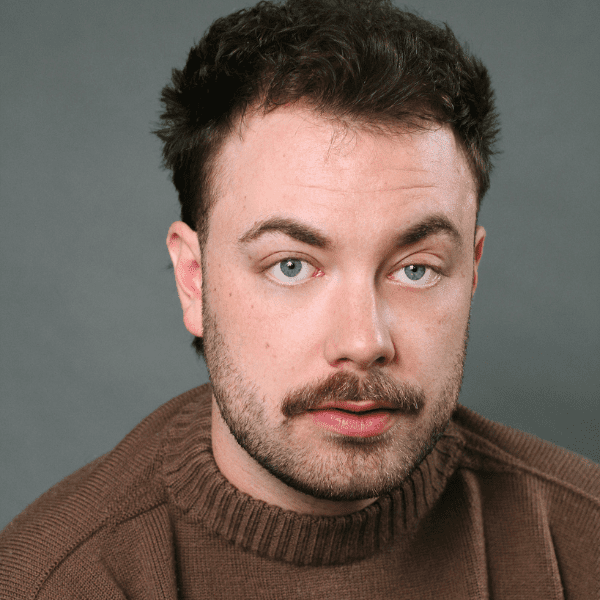 Actor Nathan Reilly in a brown sweater posed in front of a gray backdrop