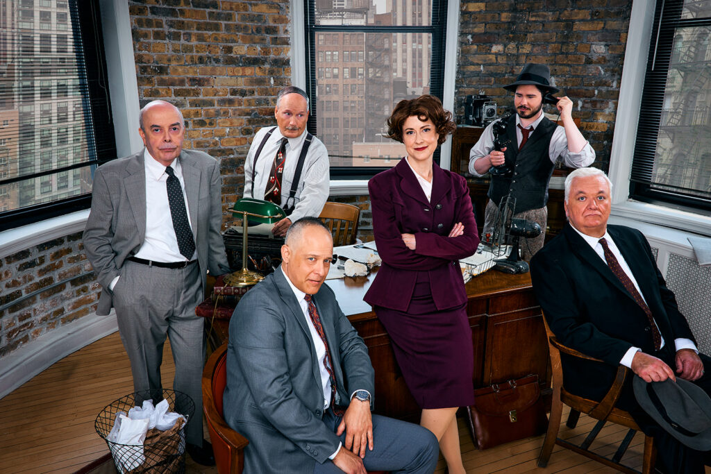 Actors Timothy Edward Kane, Kate Fry, Guy Van Swearingen, Kelan M. Smith, William Dick, and John Lister posed around a bustling news desk.