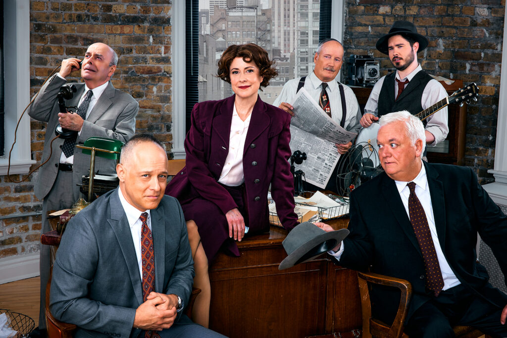 Actors Timothy Edward Kane, Kate Fry, Guy Van Swearingen, Kelan M. Smith, William Dick, and John Lister posed around a bustling news desk.