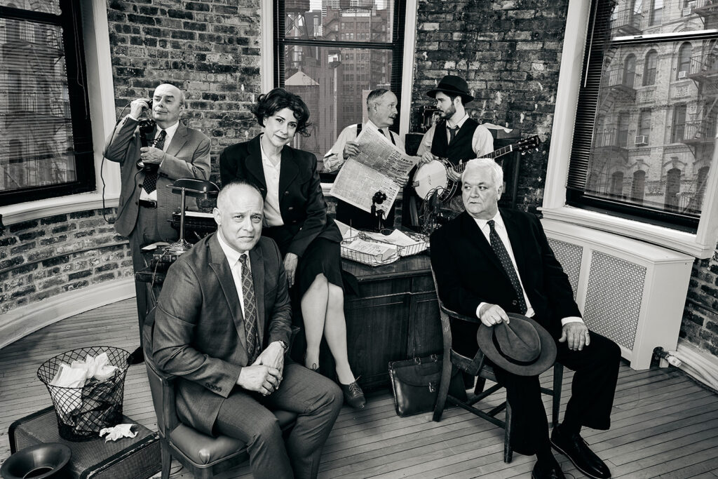 Actors Timothy Edward Kane, Kate Fry, Guy Van Swearingen, Kelan M. Smith, William Dick, and John Lister posed around a bustling news desk.