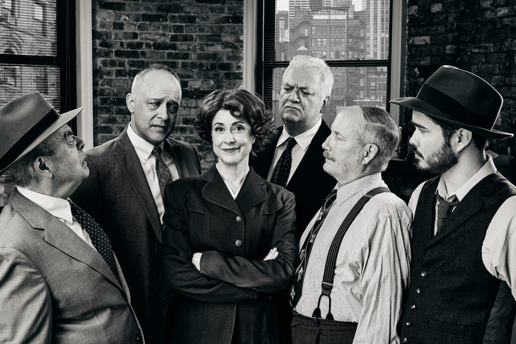 A black and white photo of Actors Timothy Edward Kane, Guy Van Swearingen, Kelan M. Smith, William Dick, and John Lister all posed around actress Kate Fry, staring her down as she smiles at the camera