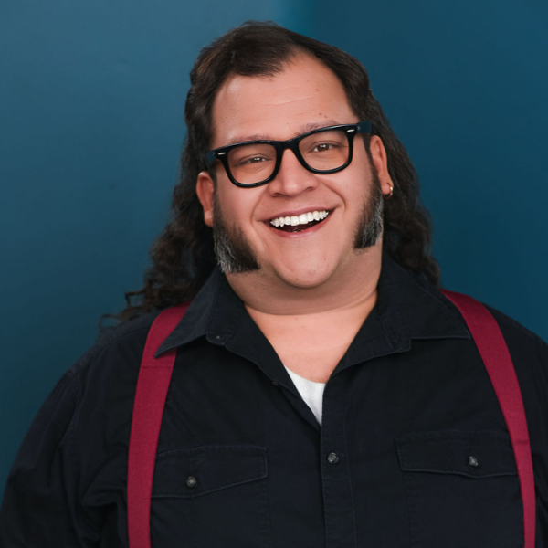 Zack _SLiM_ Simonmini Picture Zack, smiling and posed in front of a blue backdrop. He wears black framed glasses, a blue button up shirt, and red suspenders.