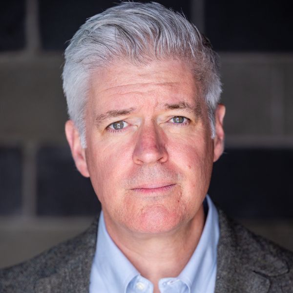 Actor Peter Moore in a blue shirt and gray jacket in front of a gray backdrop