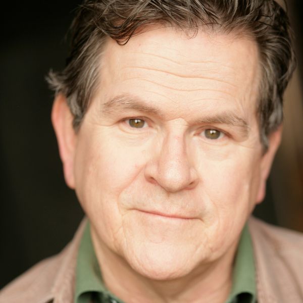 Actor Tim Kazurinksky in a brown jacket and green shirt in front of a brown backdrop