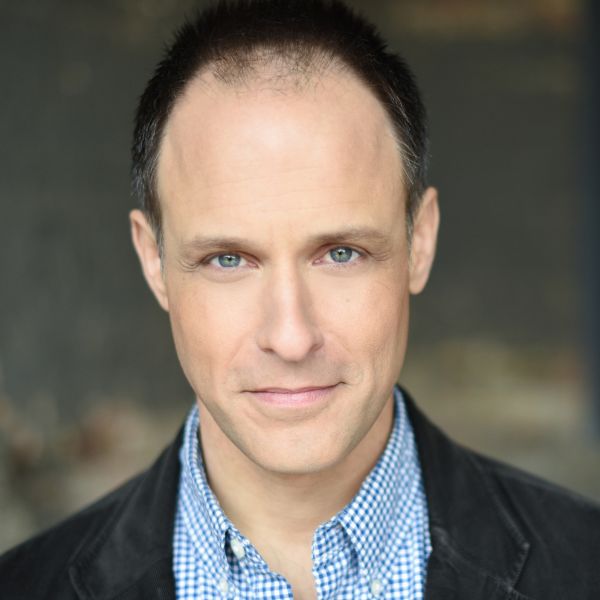 Actor Timothy Edward Kane in a blue checkered shirt and a black jacket in front of a gray backdrop