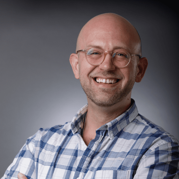 Mikey with his arms folded in a white and blue checkered shirt, posed in front of a gray backdrop